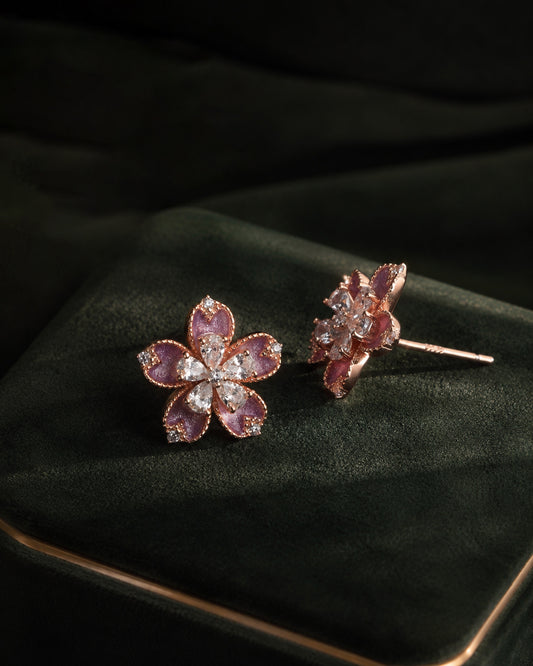 Rose gold flower-shaped stud earrings with pink enamel and white stones on a green velvet cushion