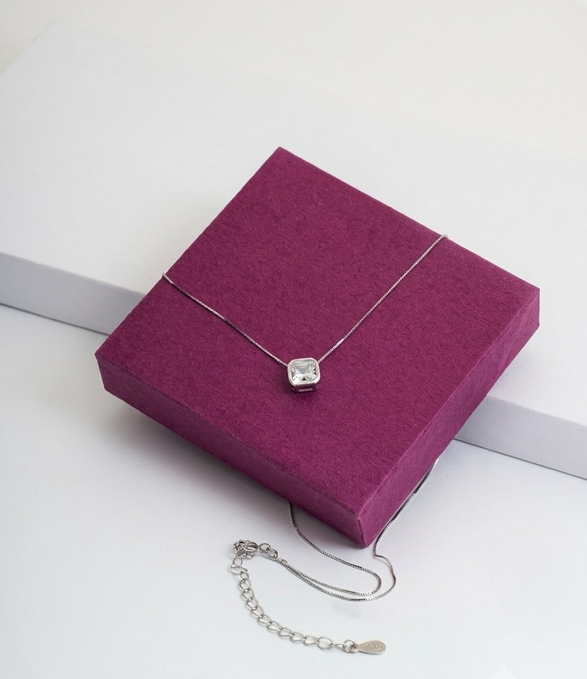 A silver necklace with a square-cut clear gemstone pendant, displayed on a textured magenta jewelry box with its chain and extender chain visible against a clean white background.