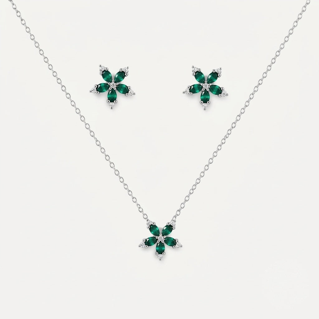 A silver jewelry set featuring a delicate chain necklace and matching stud earrings, each adorned with a five-petal flower design in polished green stones and small clear accent stones, set against a light gray background.
