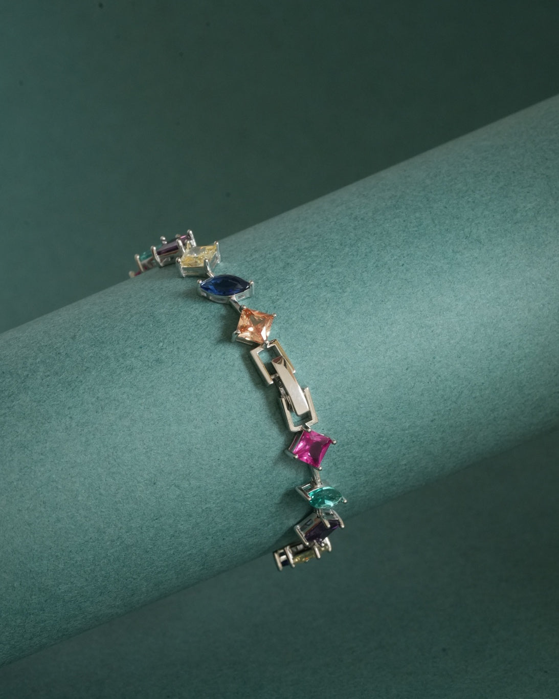 The 925 Sterling Silver Multicolor Station Bracelet draped over a minimal sculptural form, highlighting its lightweight construction and rhodium finish. On teal green background 