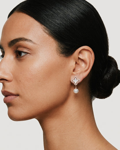 Close-up of a person wearing rose gold clover earrings with white inlay and a dangling pearl.