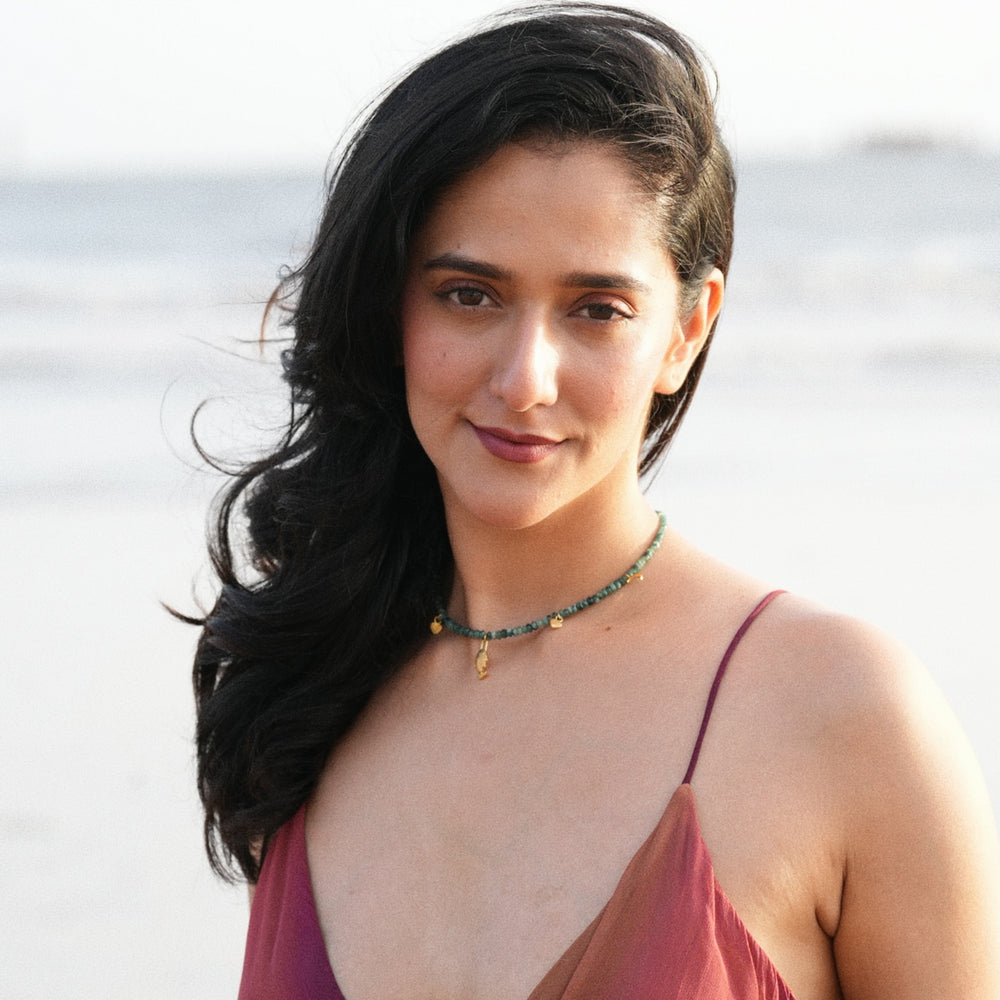A woman with long dark hair stands on a beach wearing a rust-colored spaghetti-strap top and a beaded necklace with small gold charms.