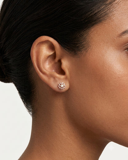 A close-up of a model's ear wearing a small flower-shaped stud earring with clear stones in a gold-toned setting, against a clean light background.