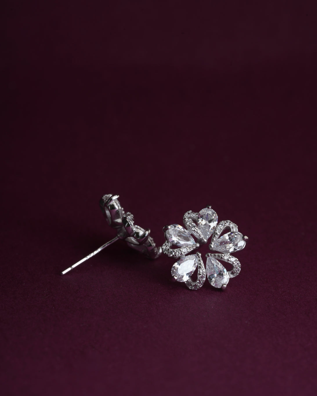 A pair of silver-toned flower stud earrings with clear teardrop-cut gemstones, displayed on a dark burgundy background.