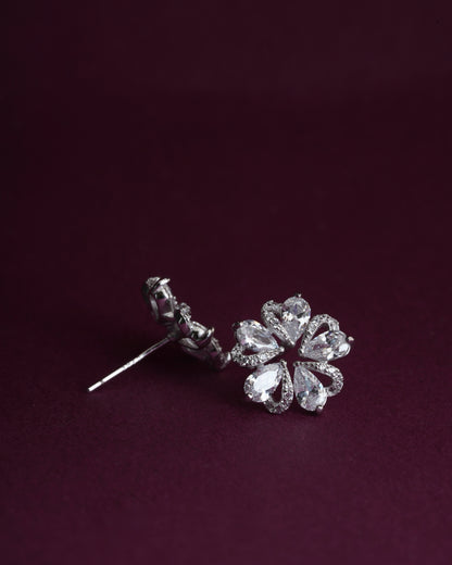 A pair of silver-toned flower stud earrings with clear teardrop-cut gemstones, displayed on a dark burgundy background.