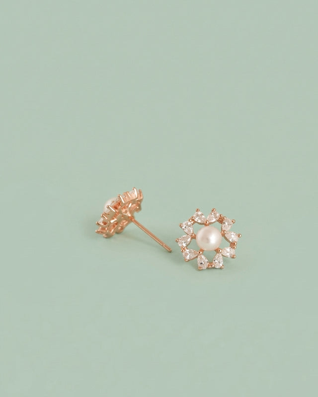 A pair of rose gold flower stud earrings, one showing the back with a screw-back post and the other showing the front with a central white pearl surrounded by small clear stones, set against a solid sage green background.