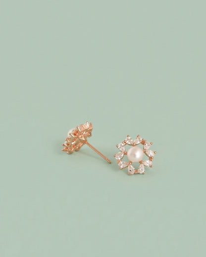 A pair of rose gold flower stud earrings, one showing the back with a screw-back post and the other showing the front with a central white pearl surrounded by small clear stones, set against a solid sage green background.