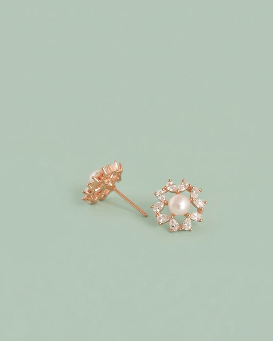A pair of rose gold flower stud earrings, one showing the back with a screw-back post and the other showing the front with a central white pearl surrounded by small clear stones, set against a solid sage green background.
