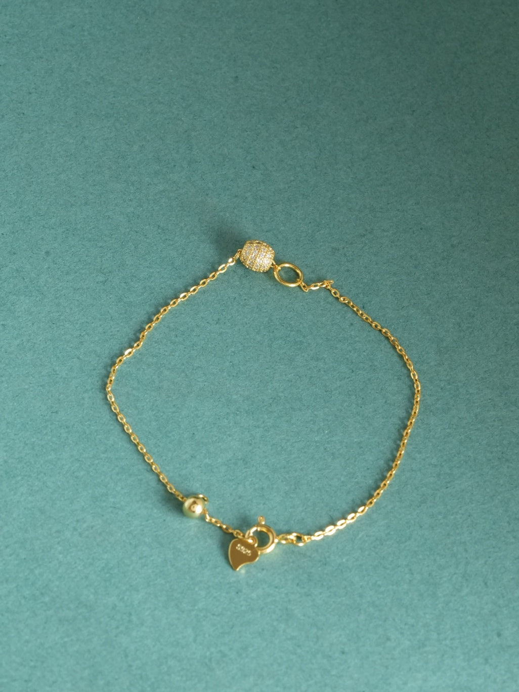 A delicate gold chain bracelet with a small heart charm engraved with 'LOVE', a tiny bead, and a textured spherical bead, displayed on a textured teal background.