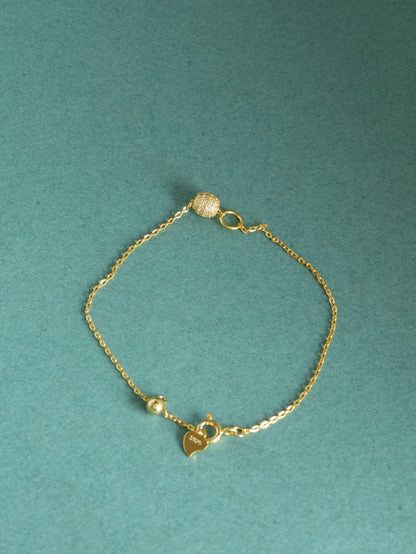 A delicate gold chain bracelet with a small heart charm engraved with 'LOVE', a tiny bead, and a textured spherical bead, displayed on a textured teal background.