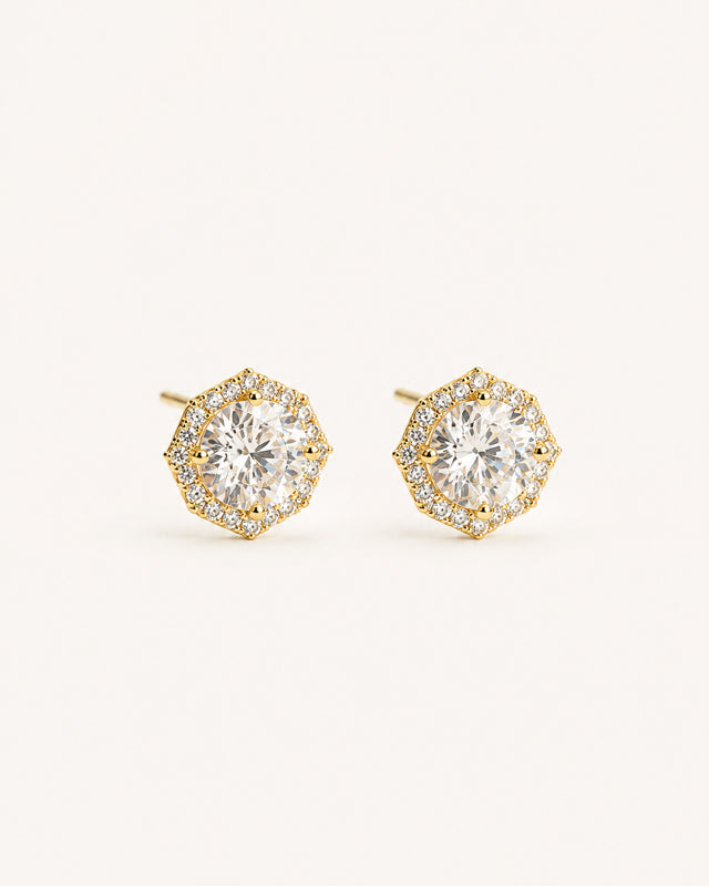 A pair of gold halo stud earrings with round clear gemstones, set against a solid light beige background.