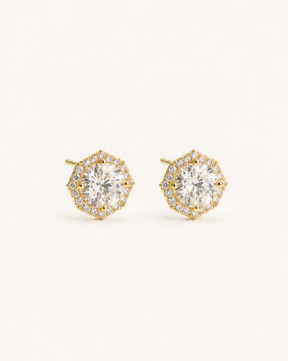 A pair of gold halo stud earrings with round clear gemstones, set against a solid light beige background.