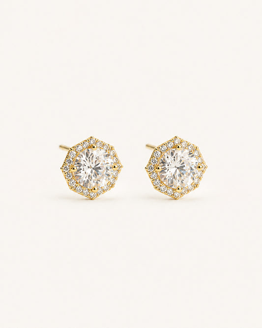 A pair of gold halo stud earrings with round clear gemstones, set against a solid light beige background.