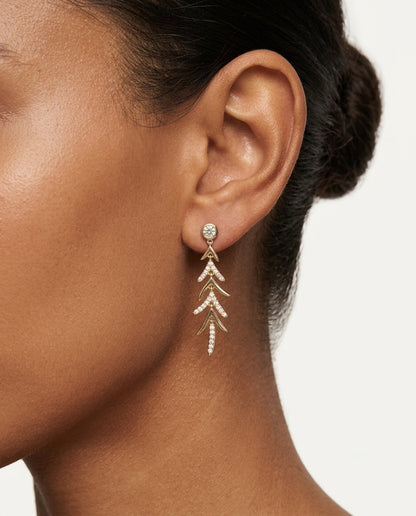 A close-up of a woman wearing a delicate gold dangle earring with a small round clear stone and layered leaf-like drops, paired with a white top.